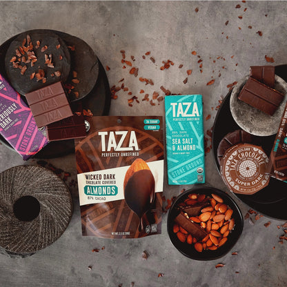 Taza dark chocolate bars, discs and chocolate covered nuts on a textured surface with various Taza packaging and ingredients.